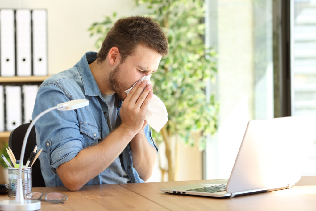 young man in an office sneezing in a tissue near a window