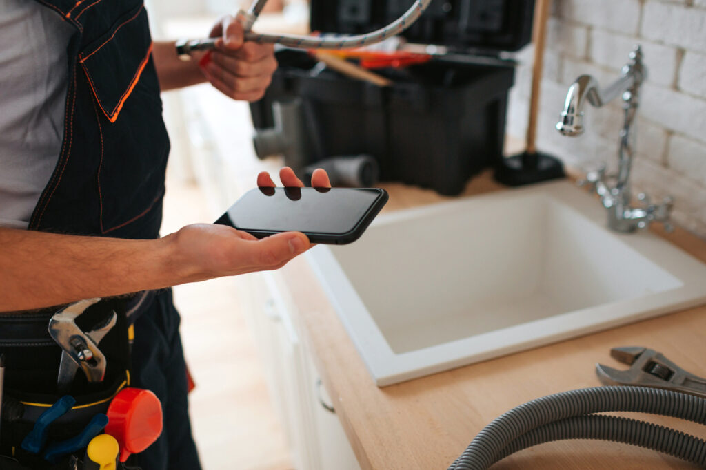 man standing in kitchen at sink holding a phone and wrench with a hose on the counter